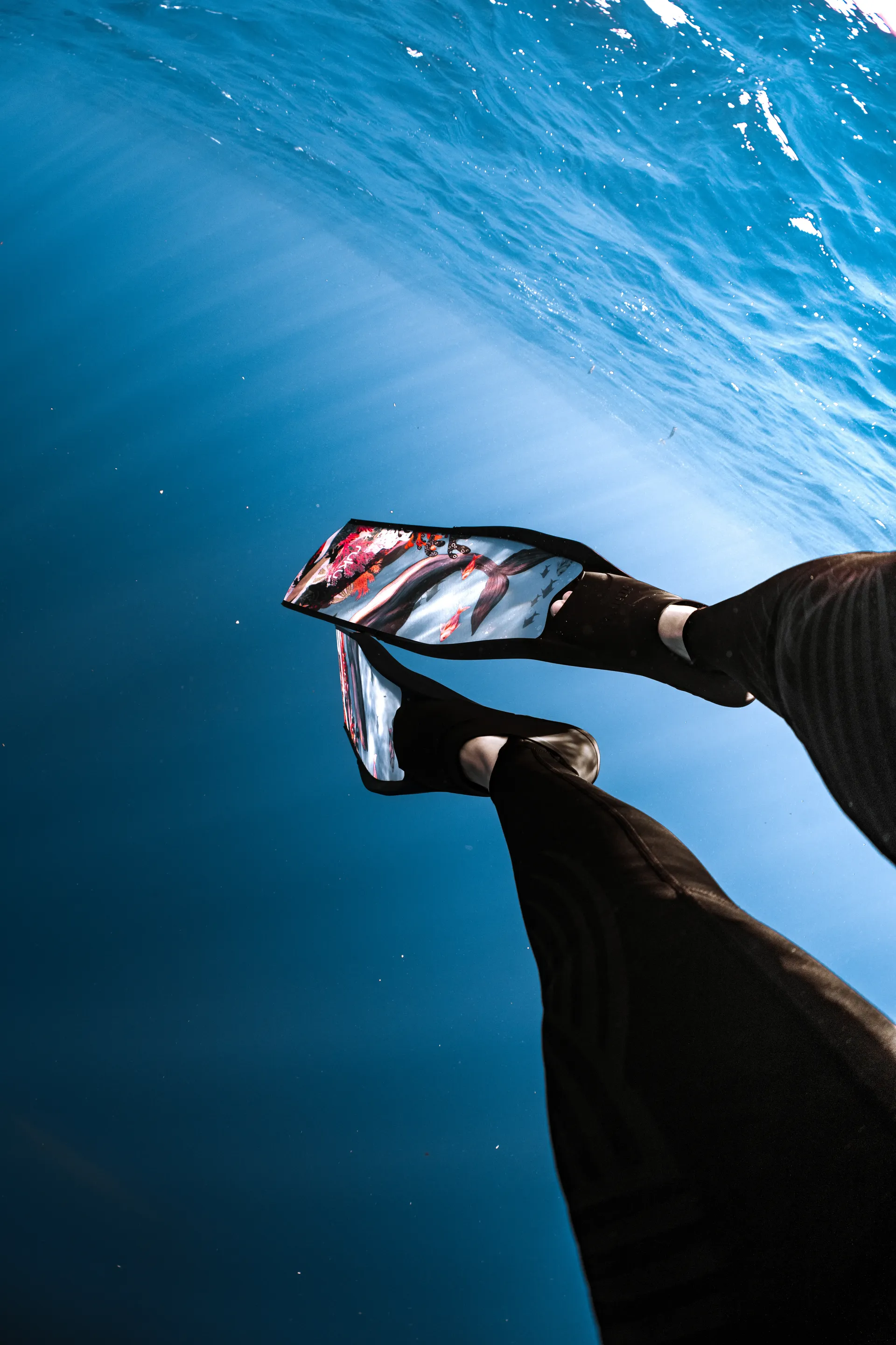 Freediving fins painted with ocean art, backlit at the surface — detail shot